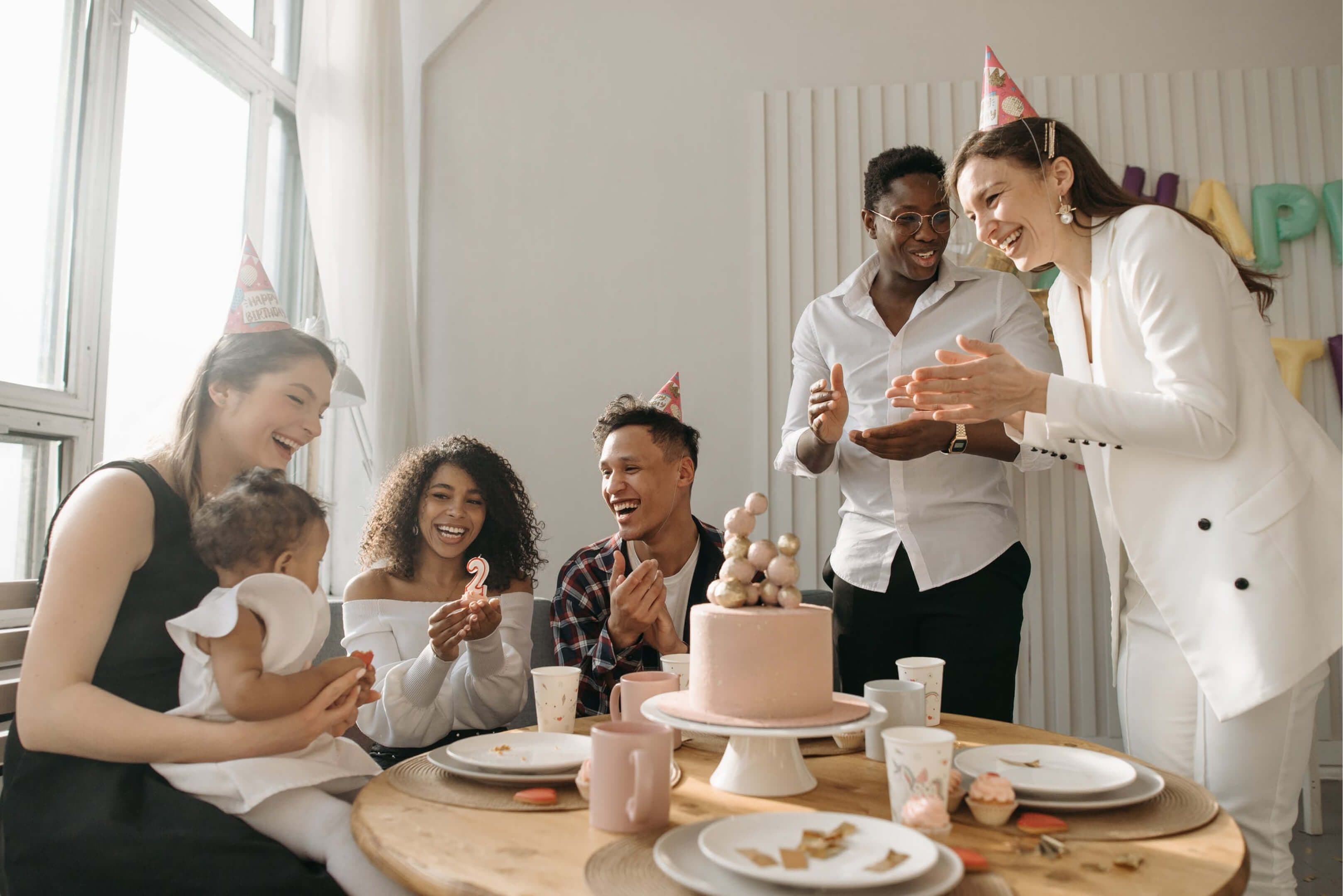 A family celebrating a toddler’s birthday, with a woman smiling and wearing a party hat while holding a child.